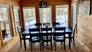 Una cabaña de madera con una mesa de comedor, sillas, una araña y ventanas amplias con vistas nevadas.