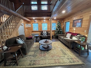 A cozy cabin interior with wooden walls, a staircase, a Christmas tree, and two sofas.
