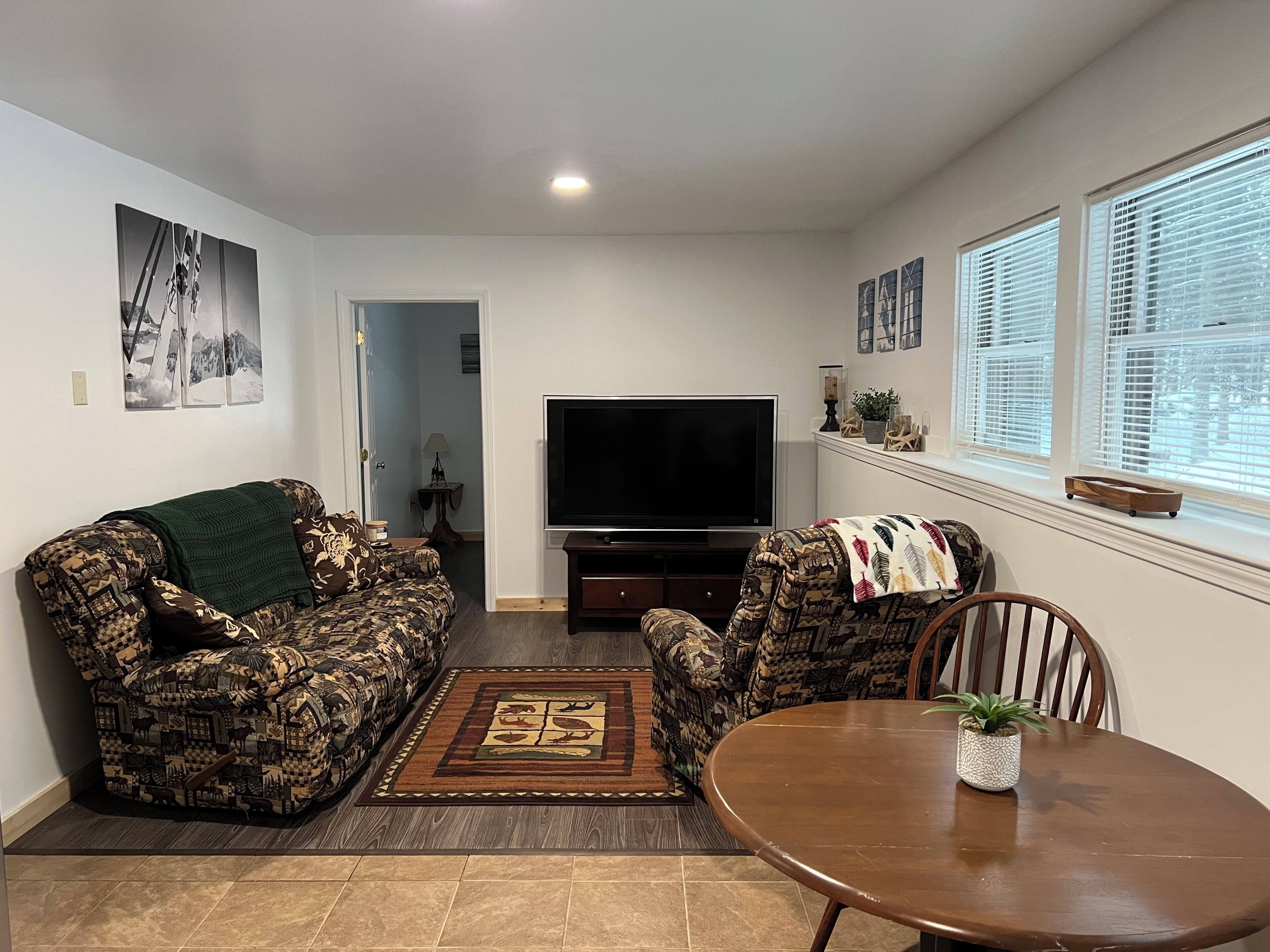 A living room with patterned sofas, a flat-screen TV, a dining table, and a window with blinds.