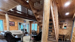 Interior de una cabaña de madera con sofá, sillas, una mesita, un tapete y una escalera.
