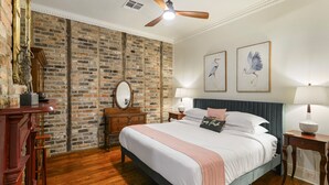 A bedroom with a brick wall, a bed with a blue headboard, two wall-mounted lamps, and a ceiling fan.