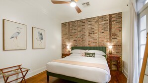 A bedroom with a brick-patterned wall, a green upholstered headboard, a wooden bed frame, a wooden bench, and two wall-mounted lamps.