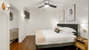 Une chambre à coucher comprenant un lit, une table de chevet, une lampe fixée au mur, un ventilateur de plafond et des œuvres d’art encadrées.