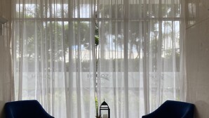 Una habitación con un amplio ventanal cubierto por cortinas de gasa, una pequeña mesa con una planta en maceta y dos sillas azules.