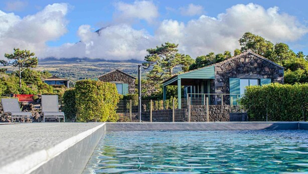 A pool area with a stone building, a covered seating area, and a view of hills in the background. - Refúgio da Montanha (Sao Roque do Pico)