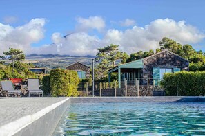 A pool area with a stone building and a covered seating area, surrounded by greenery and a mountainous backdrop. - Refúgio da Montanha (Sao Roque do Pico)