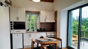A compact kitchen with white cabinets, a microwave, a coffee maker, and a sink. There's a dining table with chairs and a view of greenery through a large window. - Refúgio da Montanha (Sao Roque do Pico)