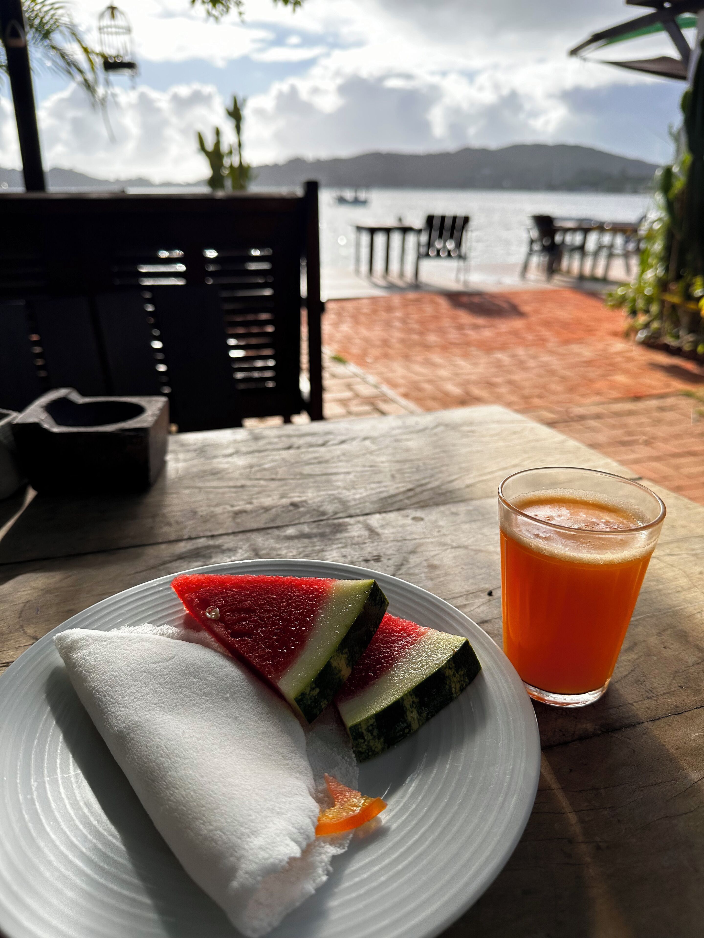 A table with a plate of food and a glass of juice, overlooking a body of water and mountains.