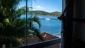 A window view of a coastal area with a boat on the water, palm trees, and a hilly landscape in the distance.