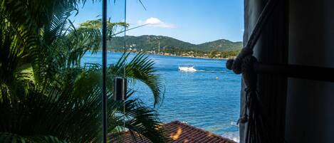 A window view of a coastal area with a boat on the water, palm trees, and a hilly landscape in the distance.