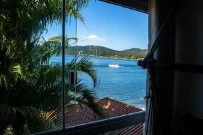 A window view of a coastal area with a boat on the water, palm trees, and a hilly landscape in the distance. - Lagoa Nômade (Florianópolis)