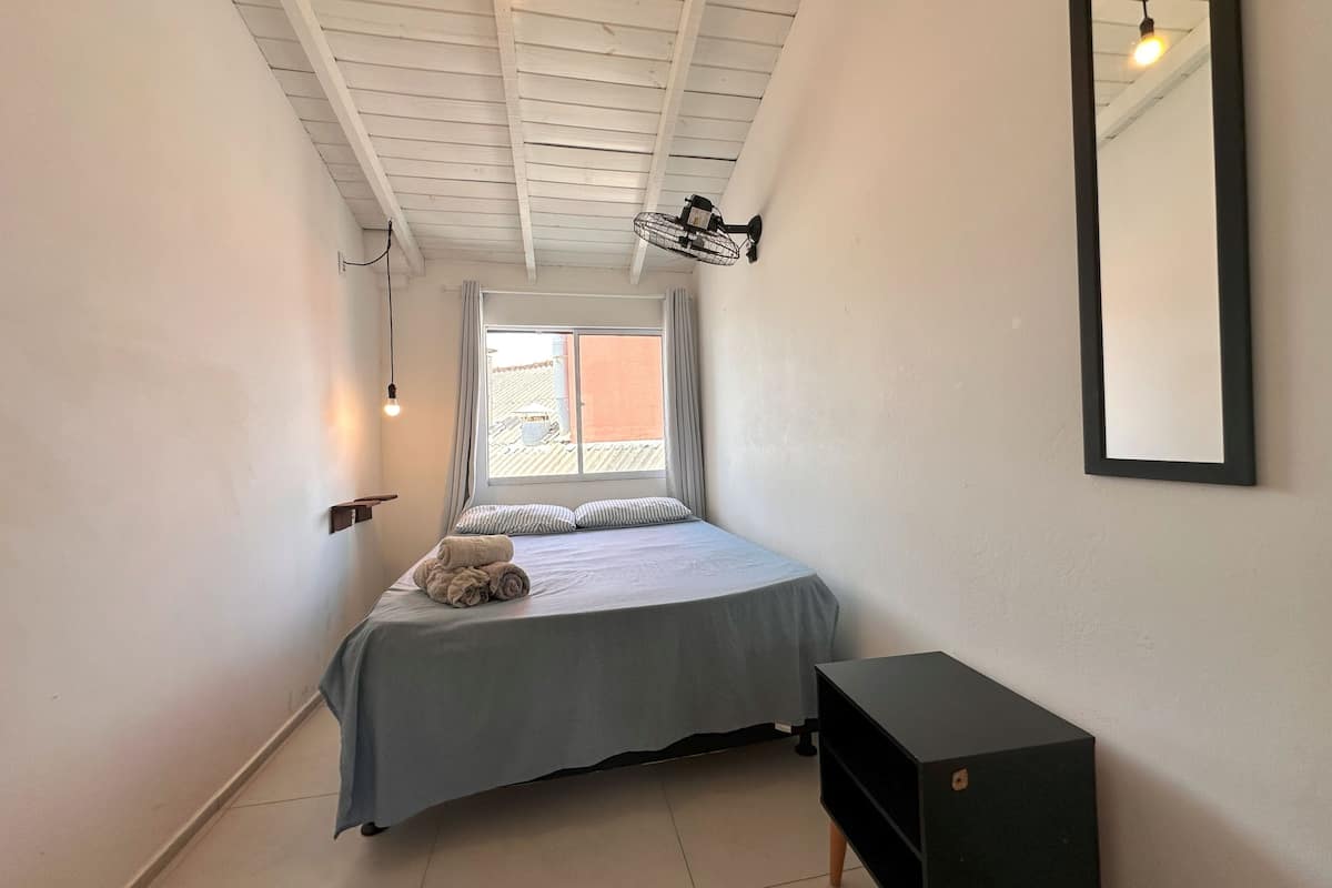 A bedroom with a bed, a small bedside table, a wall-mounted mirror, and a window with curtains.