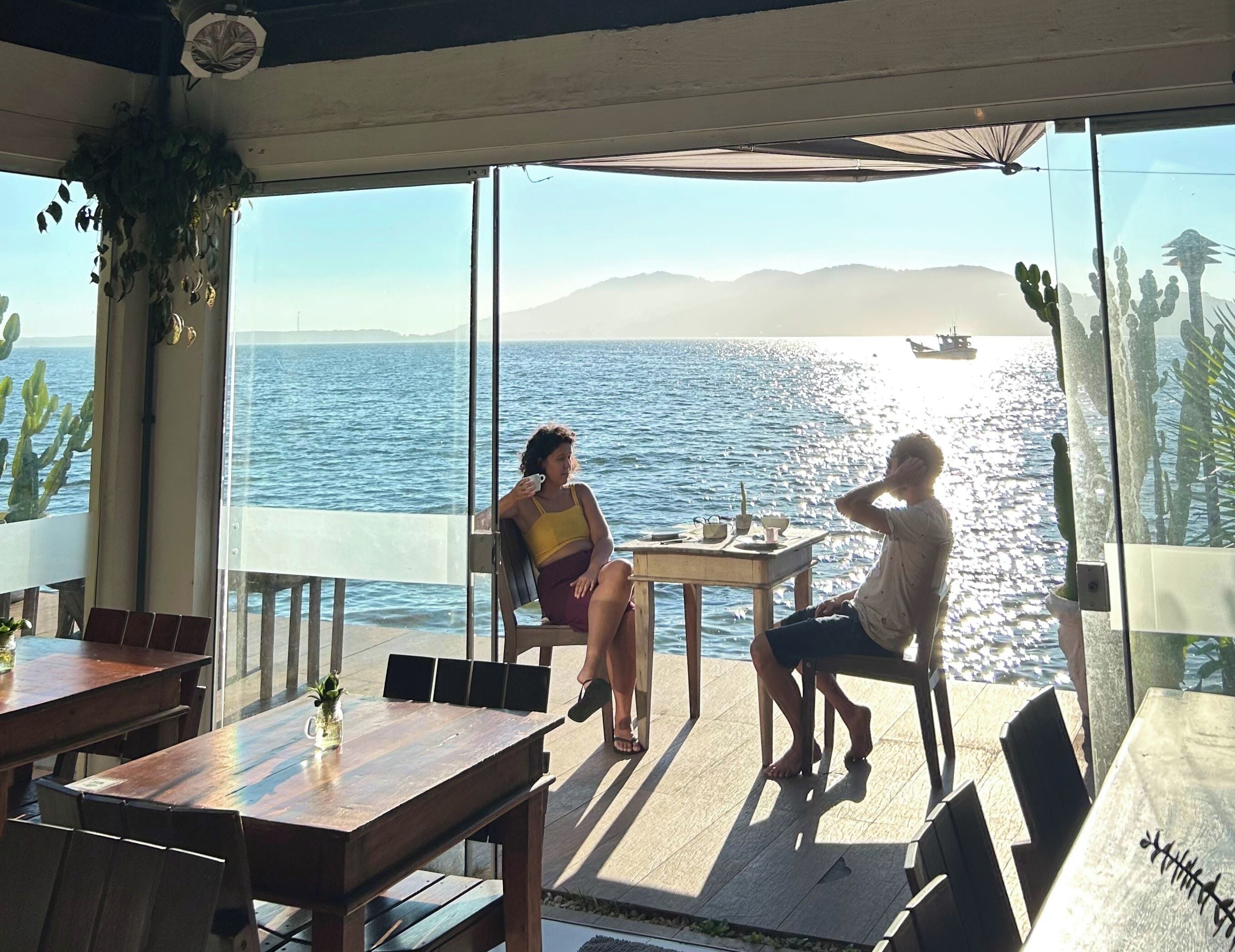 A terrace with a view of the sea, wooden tables, chairs, and a small table with a cup and saucer.