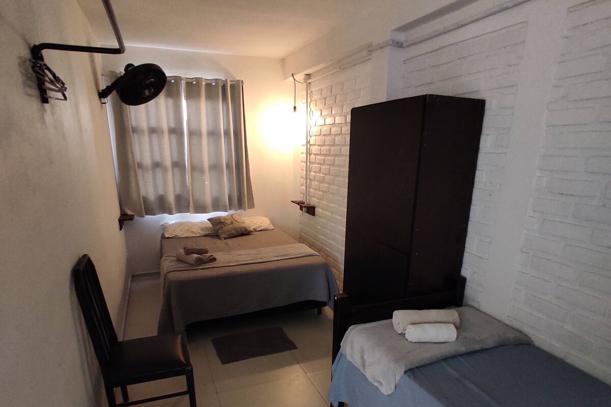 A small, compact room with a bed, a chair, a wardrobe, and a window with curtains.