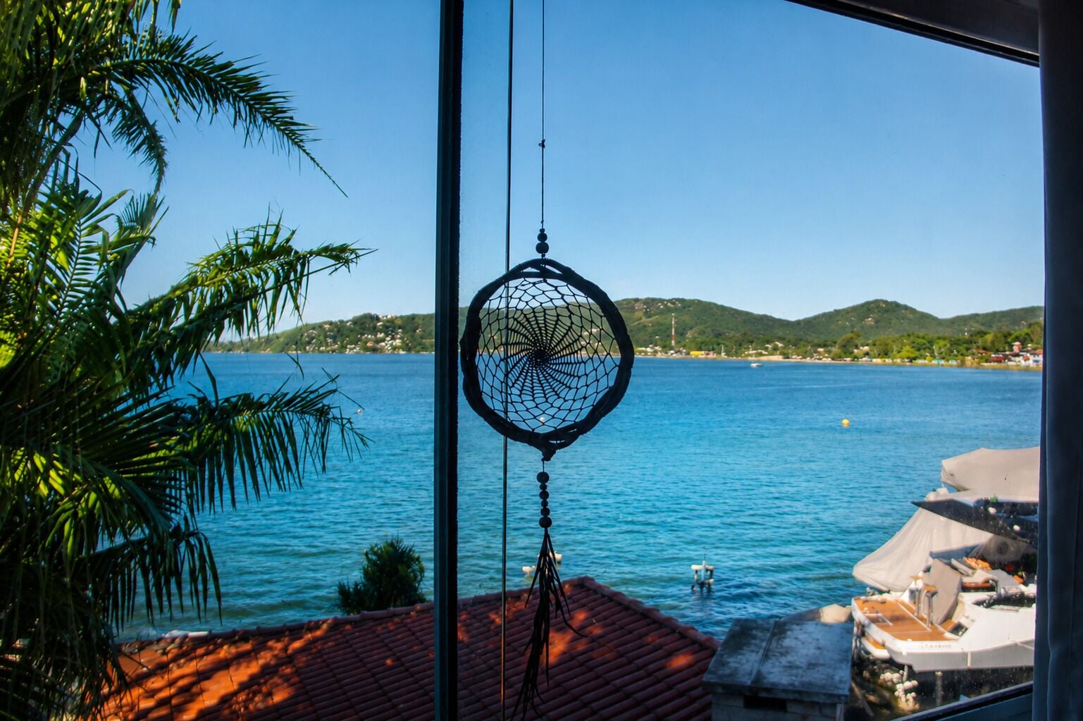 Un attrape-rêves est suspendu près d’une fenêtre donnant sur une vue côtière, avec un ciel dégagé, des palmiers et un quai pour bateaux.