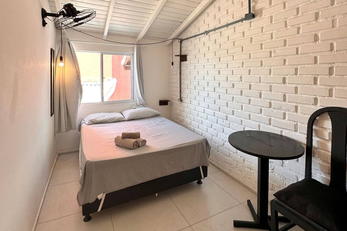 A bedroom with a bed, a small table, a chair, a window with curtains, and a brick wall.