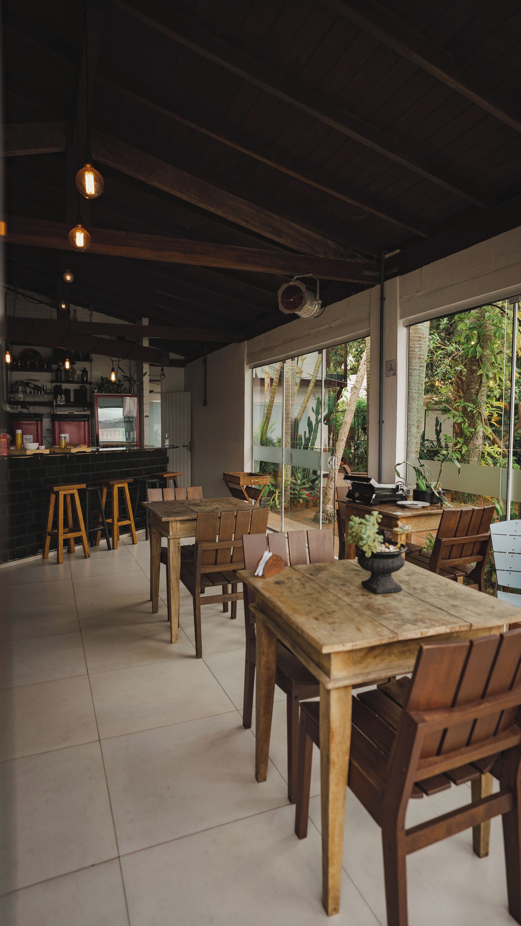 Un amplio comedor interior con mesas y sillas de madera, una zona de bar con taburetes, y grandes ventanales que ofrecen vistas al exterior.