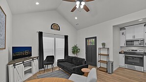 A modern living room with a black sofa, a wooden dining table, and a TV. There is a kitchen with white cabinets and a stainless steel oven. - Tiny Twin Lia, New 2 bedroom Fully Furnished Midterm in beautiful Waco (Waco)