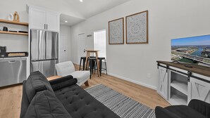 A modern kitchen with stainless steel appliances, a dining area with a white chair and bar stools, and a large flat-screen TV displaying a cityscape. - Tiny Twin Lia, New 2 bedroom Fully Furnished Midterm in beautiful Waco (Waco)