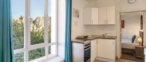 A compact kitchen with a gas stove, a sink, and cabinets. There is a window with blue curtains, a clock on the wall, and a bed in the adjacent room.