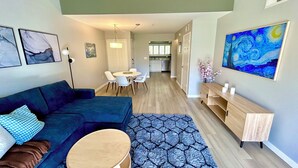 Un saló modern amb un sofà blau, una taula de centre rodona i una catifa estampada. Hi ha una zona de menjador amb una taula i cadires blanques, i una cuina al fons.