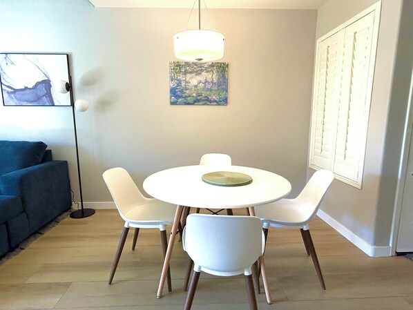 A dining area with a round white table, four white chairs, a painting on the wall, and a blue couch in the background. - Modern Condo in Prime Location - Heated Pool, Bars, Dining Nearby (Scottsdale)