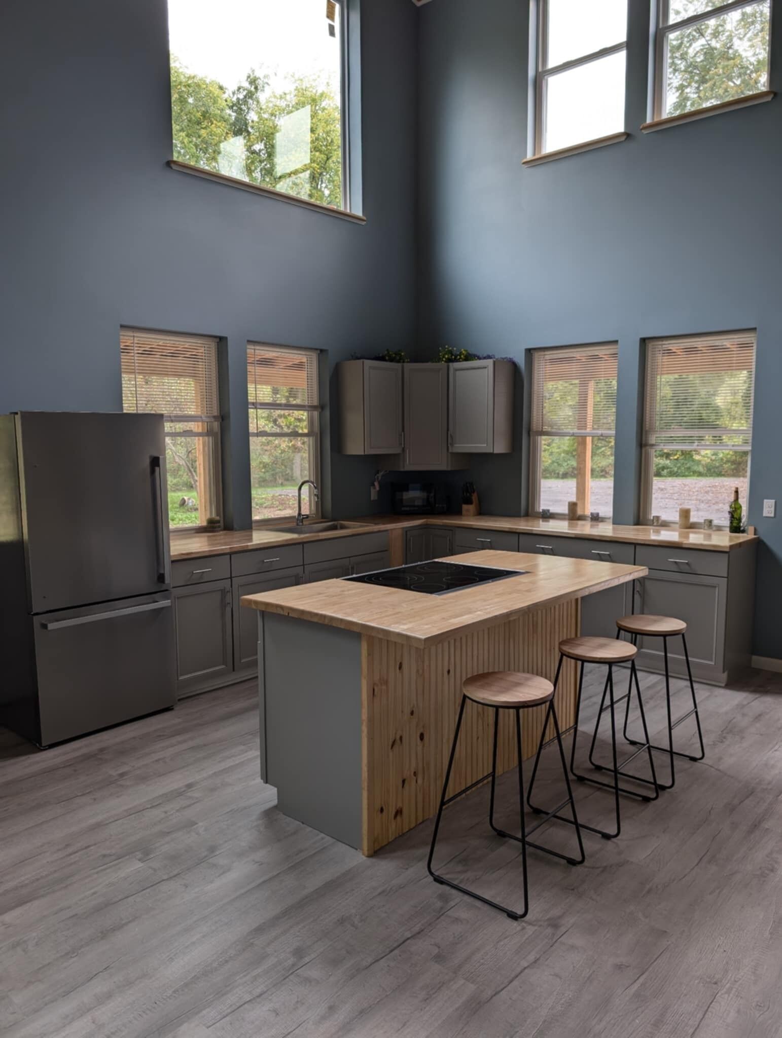 Une cuisine moderne avec des meubles gris, un îlot central en bois, des tabourets de bar et de grandes fenêtres.