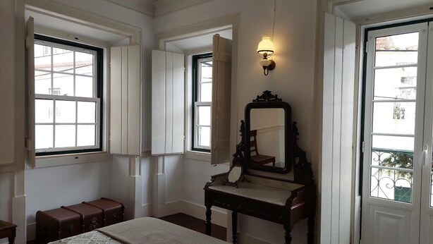 Quarto com cama, cômoda, espelho e duas janelas.
