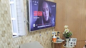 Un salon avec un téléviseur à écran plat fixé au mur, un fauteuil gris, une petite table sur laquelle est posé un vase de fleurs et un panneau décoratif mural.