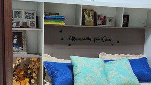 Une chambre à coucher avec un lit, une étagère remplie de livres et de photos, un vase décoratif et un mur orné d’une citation religieuse.