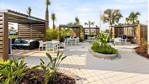 Un patio al aire libre con espacios de estar, incluyendo sofás y mesas, rodeado de palmeras y plantas.