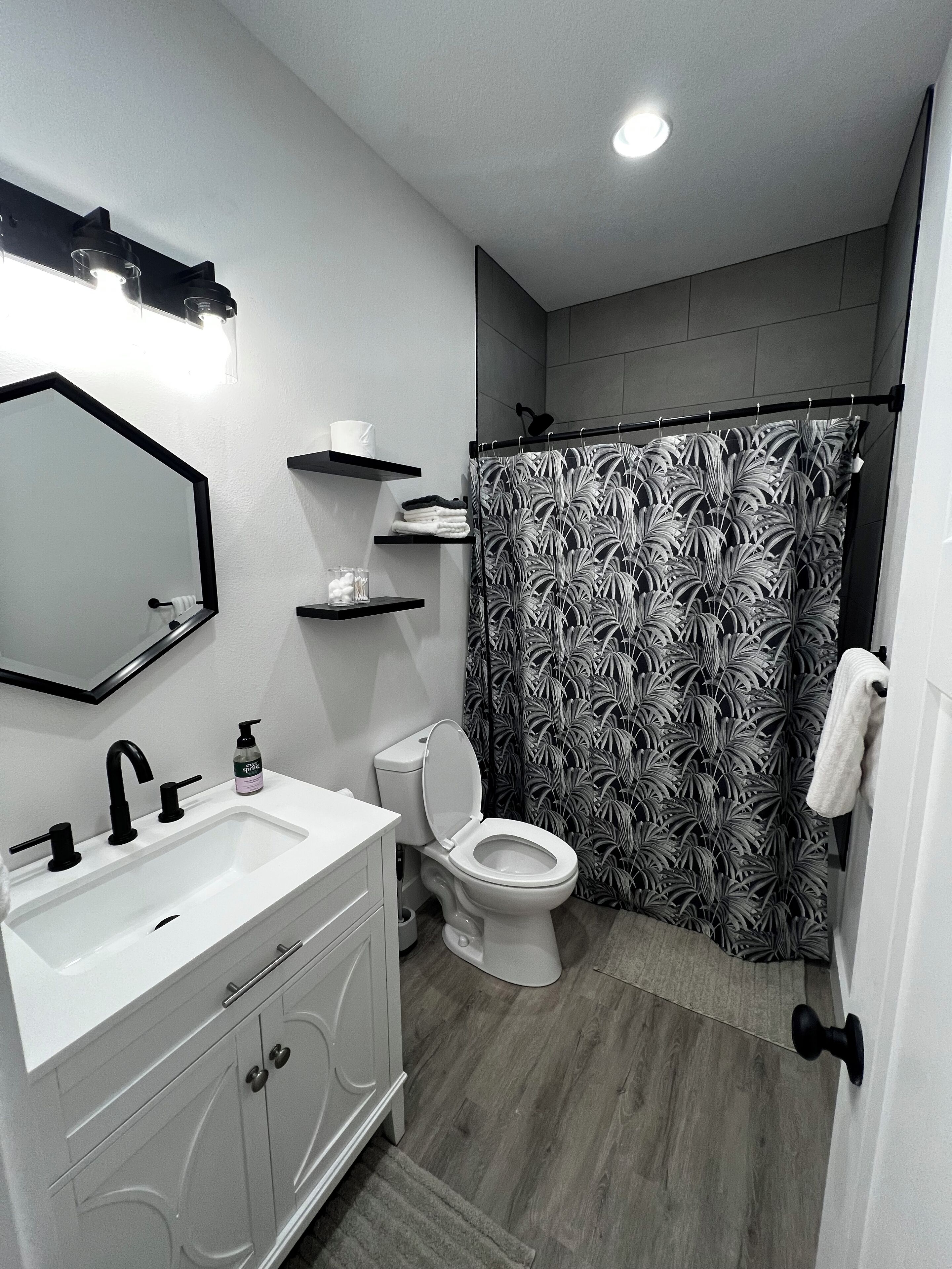 A modern bathroom with a white sink, black faucet, and a toilet. There are shelves above the toilet and a shower curtain with a leaf pattern.