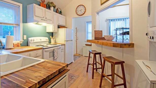 Eine Küche mit weißen Schränken, einer Holzausziehtafel, einem Herd und einem Spülbecken. Zwei Holzhocker und eine Uhr sind an der Wand angebracht.