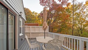 Eine Holzterrasse mit Tisch und Stühlen, ein weißer Zaun und Blick auf Herbstbäume.
