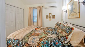 Ein Schlafzimmer mit floralem Bettbezug, einem Fellüberwurf und einem Fenster mit Vorhängen. An der Wand sind Wandleuchten und gerahmte Bilder angebracht.