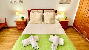 Un lit bien fait, recouvert d’une couette verte, avec deux oreillers et deux serviettes enroulées, posé sur un plancher en bois. Il y a deux tables de chevet avec des lampes et une petite plante sur chacune. La pièce a des murs blancs et une porte en bois.