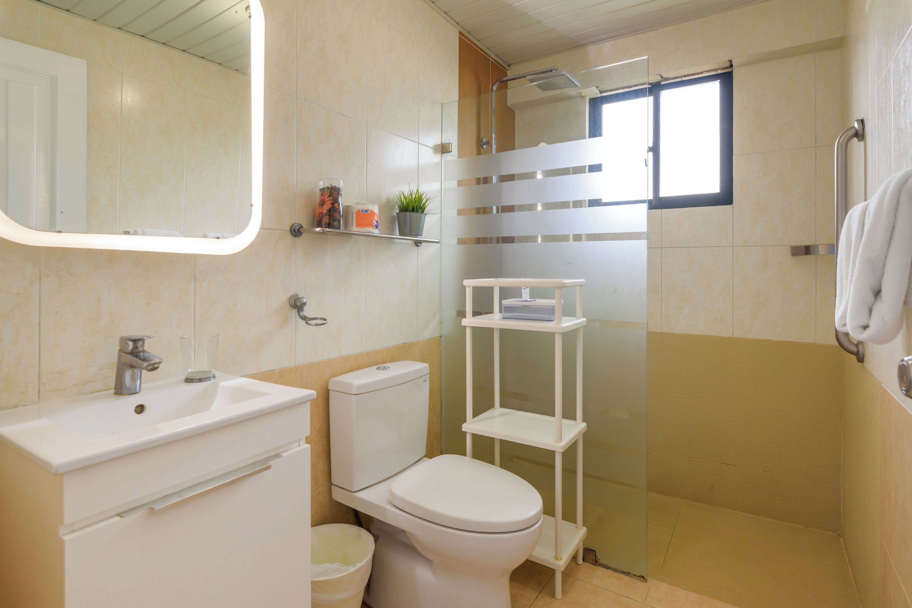 A bathroom with a toilet, sink, and mirror. There is a window providing natural light and a shower area.