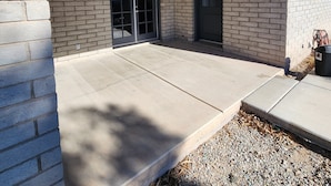A covered patio area with a concrete floor, brick walls, and a black door.