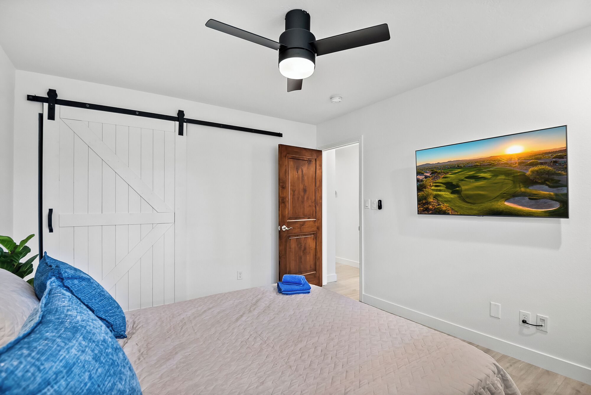 Ein Schlafzimmer mit einem Bett, einem Deckenventilator, einer Holztür, einem Wand-Fernseher und einem Landschaftsbild.