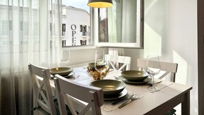 Een eettafel met witte stoelen, een hanglamp en uitzicht op gebouwen door het raam.