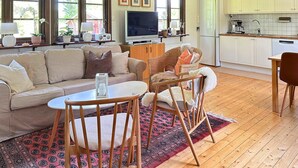 A living room with a sofa, a TV, a dining table with chairs, a kitchen area, and a window with a view of trees.