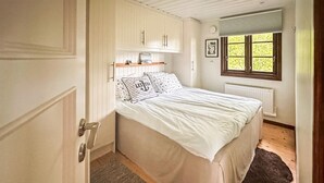 A bedroom with a bed, white walls, wooden flooring, a window with blinds, and a radiator.