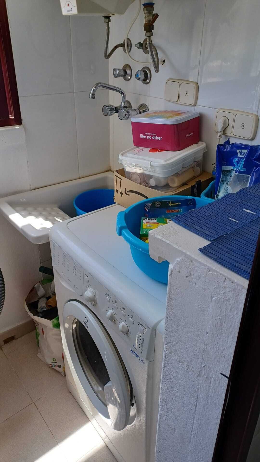 A compact kitchen with a washing machine, a sink with multiple faucets, and various containers and bags on the counter.