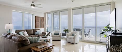 A spacious living room with a large sofa, two armchairs, a coffee table, and a TV. There are ceiling fans, large windows, and a balcony with a view.
