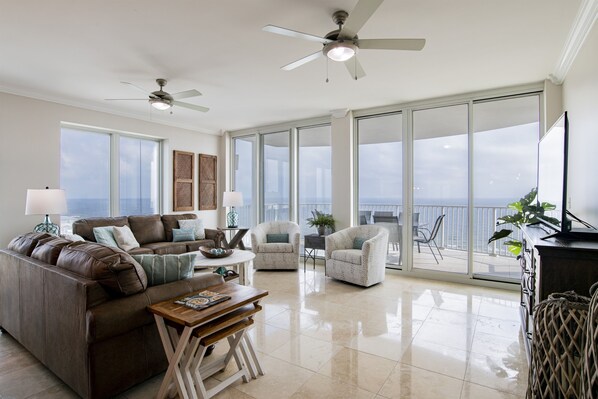 A spacious living room with a large sofa, two armchairs, a coffee table, and a TV. There are ceiling fans, large windows, and a balcony with a view.