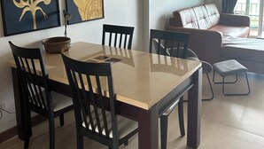 Ein Essbereich mit einem rechteckigen Tisch und vier Stühlen, einer braunen Couch und einem kleinen Hocker. Über dem Tisch hängen zwei Wandbilder.