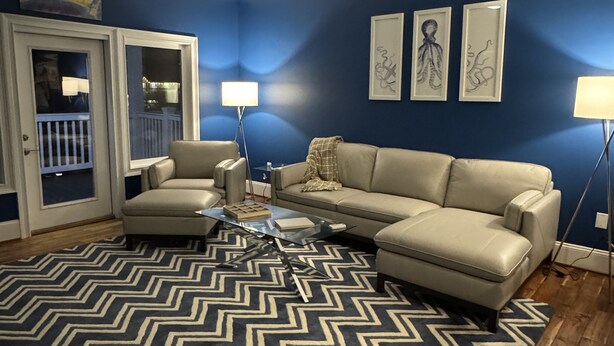 Ein Wohnzimmer mit blauer Wand, Deckenventilator, einem beigefarbenen Ecksofa, einem Glastisch und einem Teppich mit Muster.
