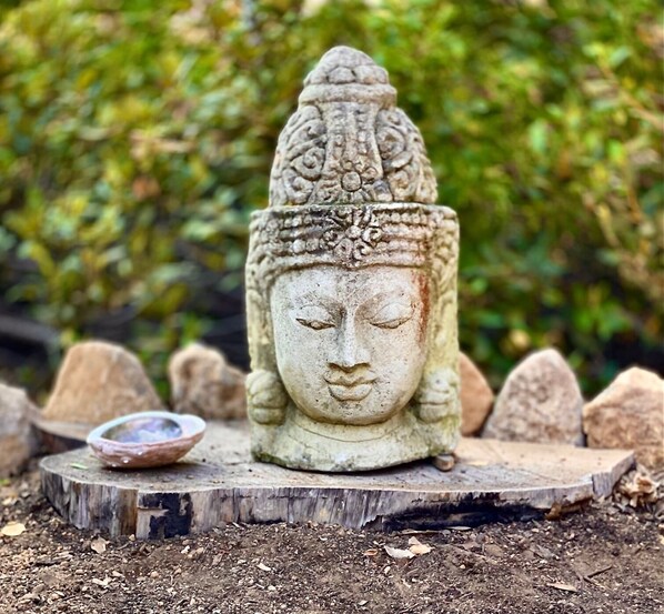 A stone Buddha head statue on a wooden platform surrounded by rocks, with a small bowl nearby.