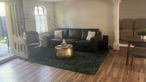 A living room with a dark leather sofa, a round coffee table, and a dark green rug. There is a window with curtains, a door with a sign, and a small table with a plant.