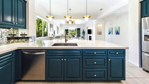 Een moderne keuken met donkerblauwe kastjes, een RVS vaatwasser en een groot eiland met een spoelbak. Boven het eiland hangen hanglampen en er is zicht op de eetruimte.